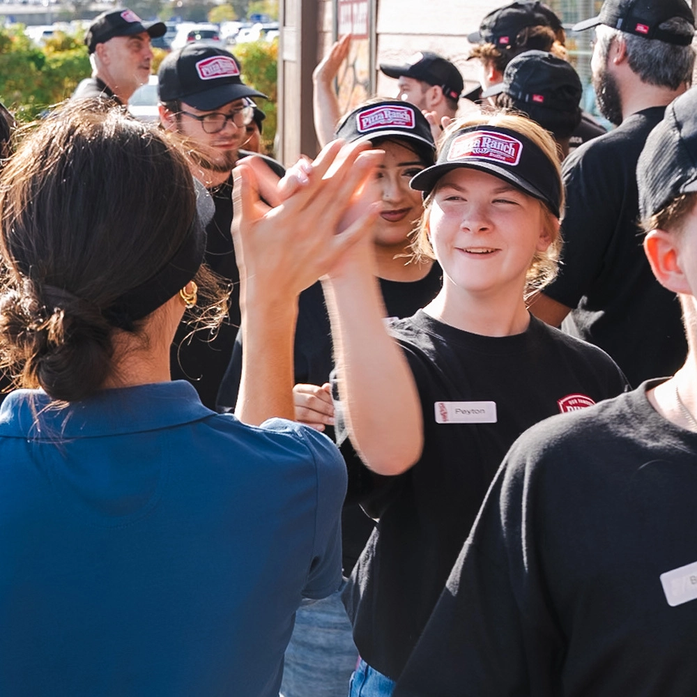 A group of people in black uniforms and caps, some with name tags, gather outdoors. They are smiling and high-fiving, celebrating together.