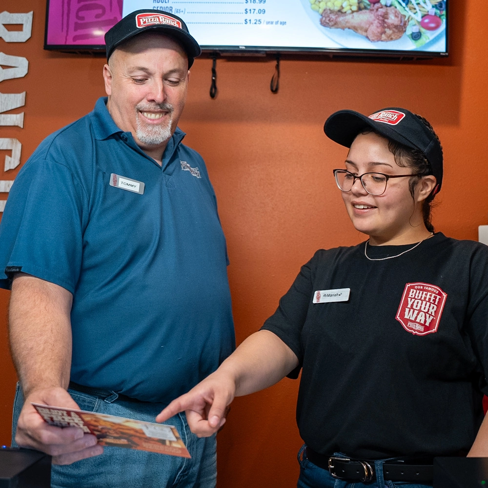 A man and a woman in uniforms smiling, with the woman pointing at a menu or flyer, standing inside a restaurant with orange walls and a digital menu board.