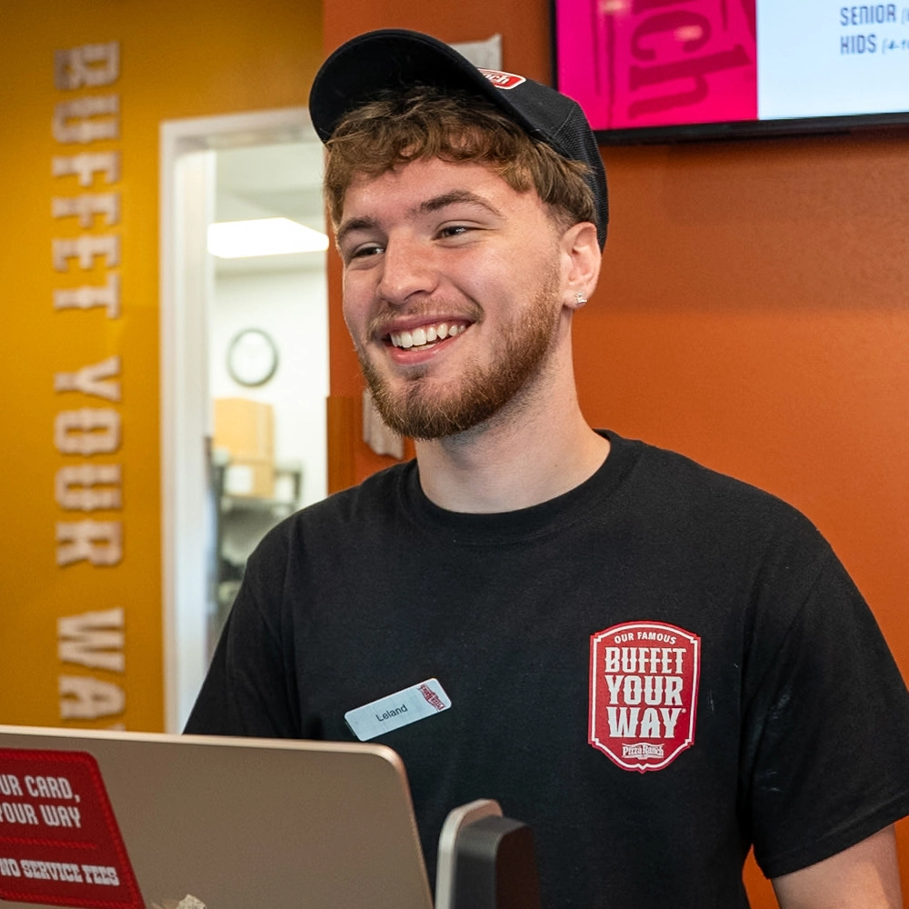 Smiling employee in a black uniform and cap, working at a computer in a Pizza Ranch restaurant with orange walls and a digital menu board behind.