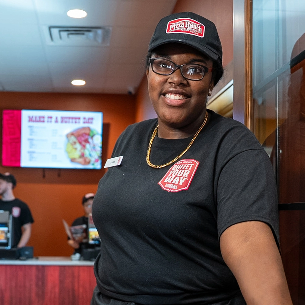 A smiling employee wearing a black Pollock's Deli T-shirt, a black cap with Pizza Ranch logo, glasses, and a gold chain, stands inside a restaurant.