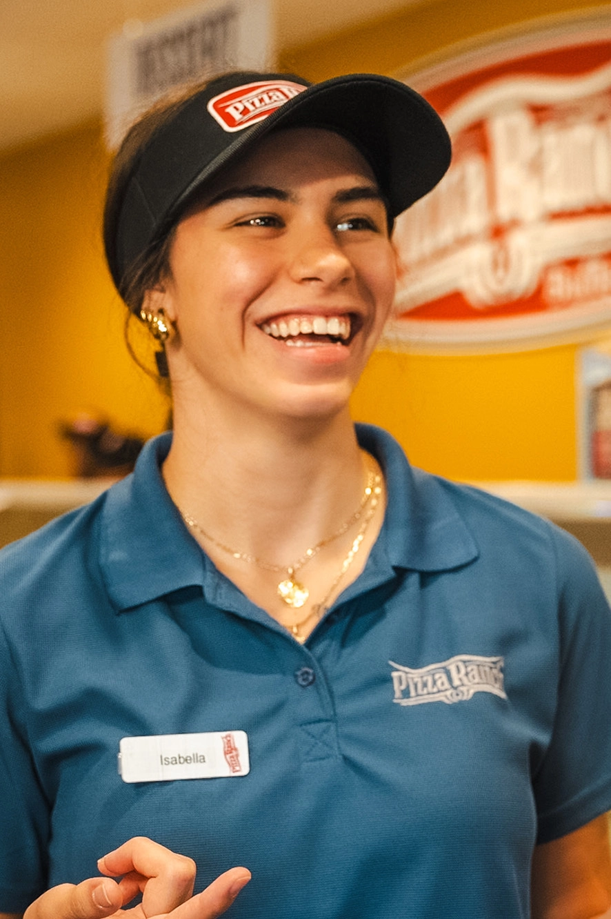 A woman wearing a blue Pizza Ranch uniform with a name tag, smiling in a restaurant. She has gold jewelry and is wearing a black visor with a Pizza Ranch logo.