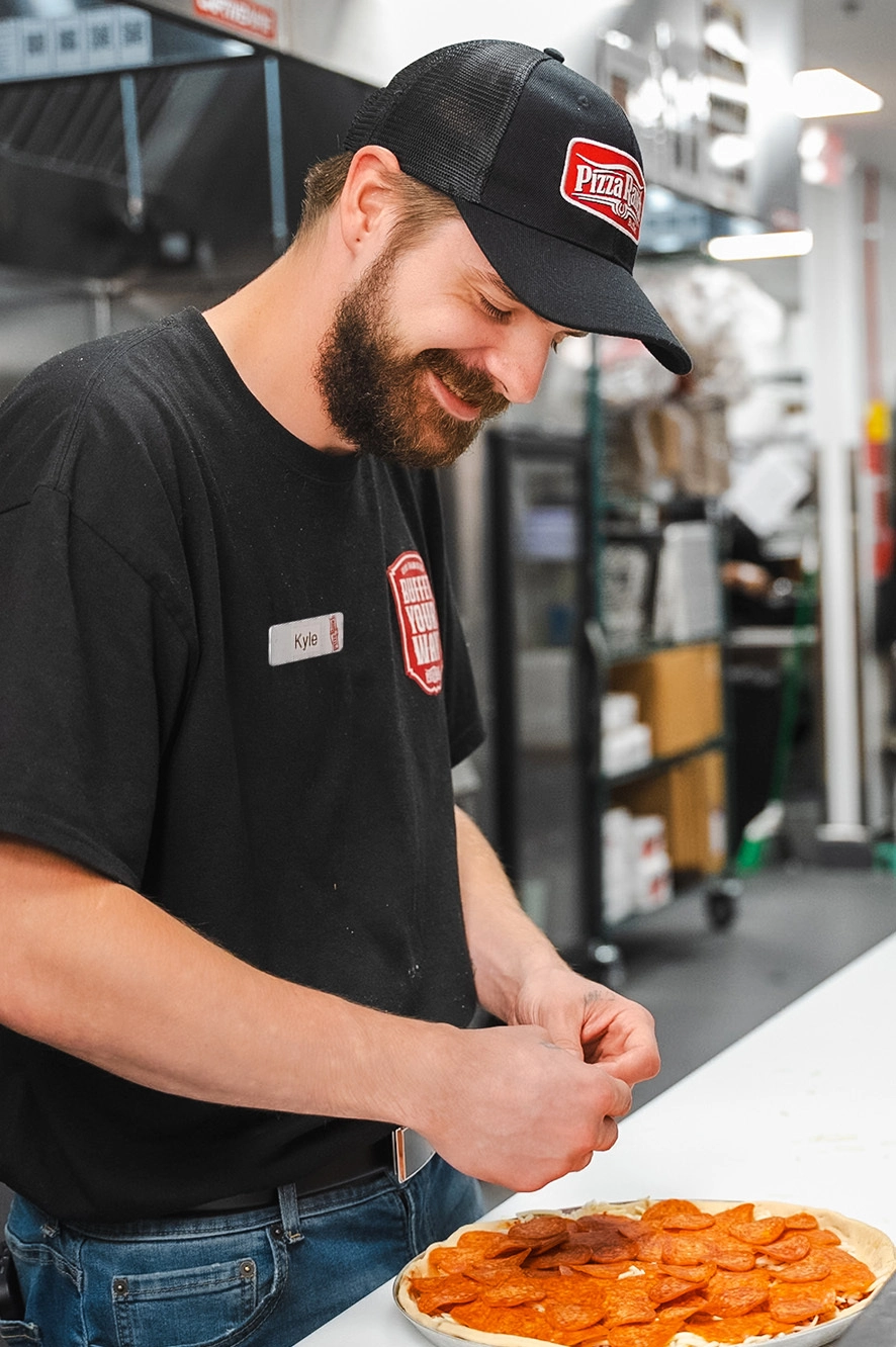A man with a beard wearing a black Pizza Ranch cap and shirt, smiling while preparing a pizza with pepperoni and sausage in a restaurant kitchen.