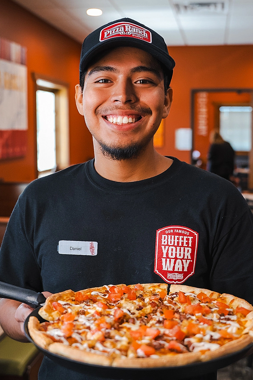 Smiling employee in a black Pizza Ranch uniform holding a freshly baked pizza inside a restaurant with orange walls.