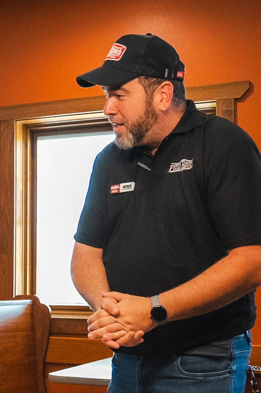 A man with a beard and earring is wearing a black uniform with a Pizza Ranch logo and a black cap, standing in a warmly lit restaurant near a window.