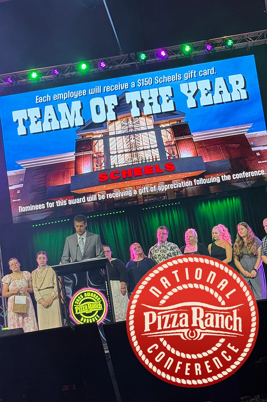 A group of people stand on a stage behind a speaker at an awards event, with a large screen above displaying "TEAM OF THE YEAR" and a gift card.