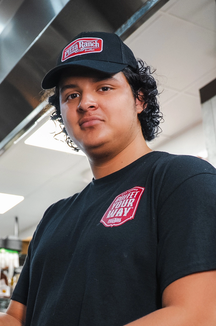 A young man with curly dark hair wearing a black Pizza Ranch cap and T-shirt, standing in a restaurant kitchen.