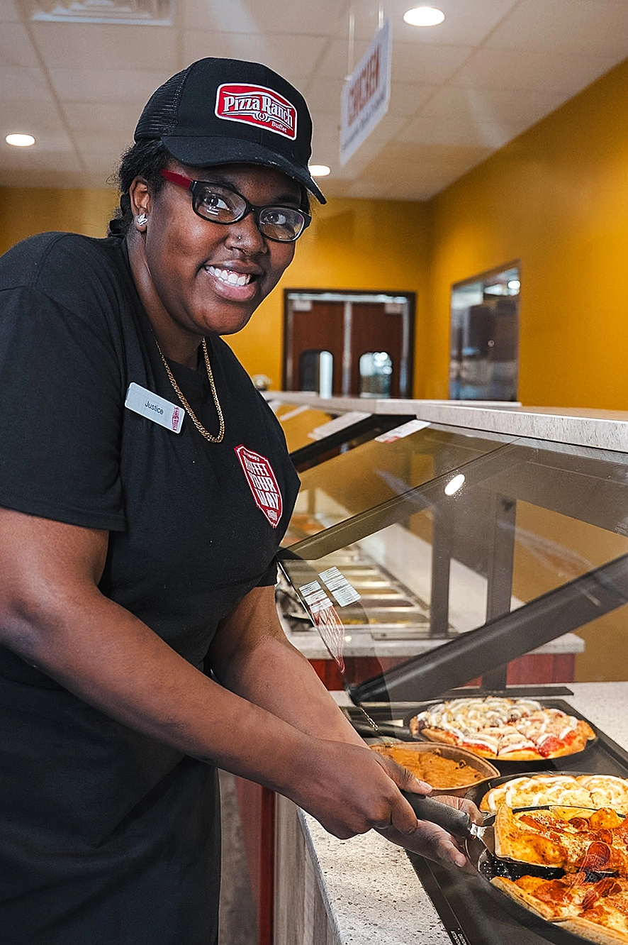 A woman wearing glasses, a black Pizza Ranch cap, and a black uniform with a name tag and logo, is serving pizza from a display case in a fast food restaurant.