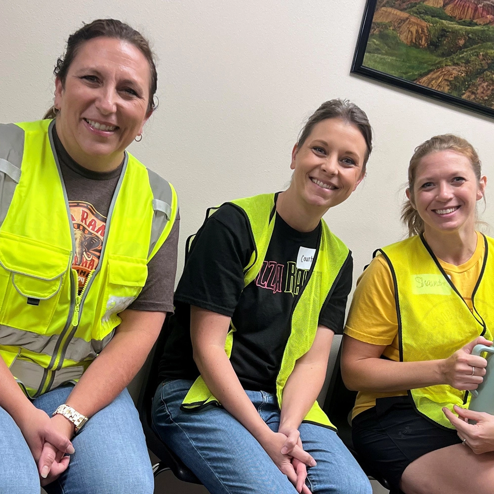 Three women sitting close together, smiling, wearing high-visibility safety vests, in an office with a landscape painting on the wall.