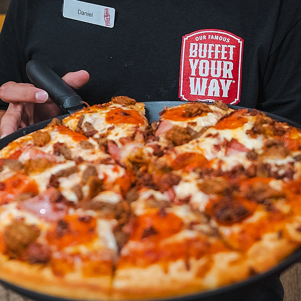 Person holding a black pizza pan with a freshly baked pizza topped with sausage, ham, and cheese, wearing a black shirt with a red and white Buffet Your Way logo.