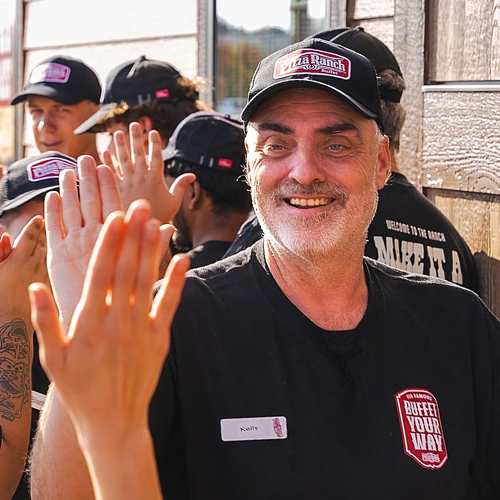 A smiling man in a Pizza Ranch cap and black shirt giving a high-five to a person whose hand is in the foreground, with others in similar caps in the background.