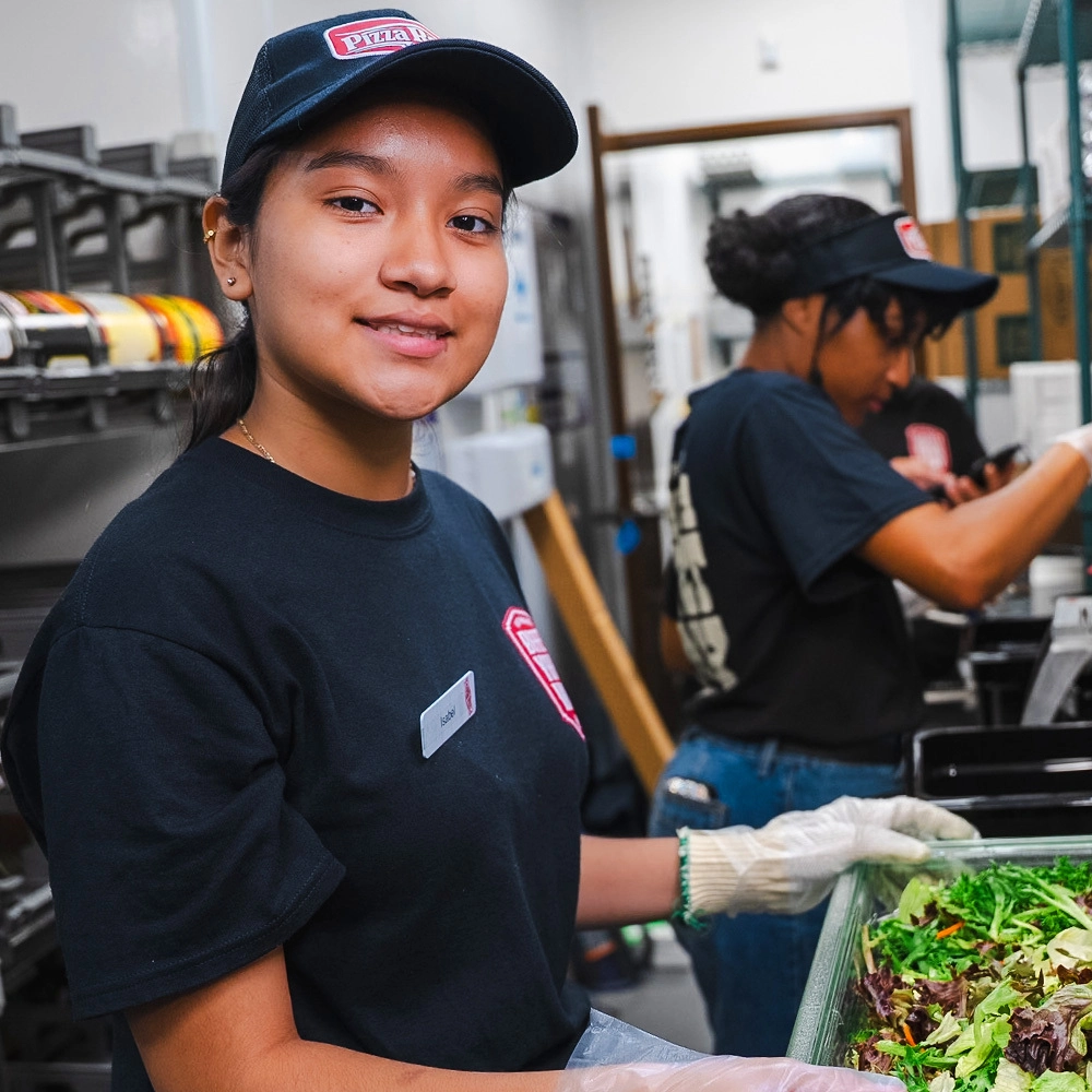 A young woman in a black Pizza Ranch uniform and cap holds a container of fresh salad while smiling at the camera, with another worker blurred in the background.