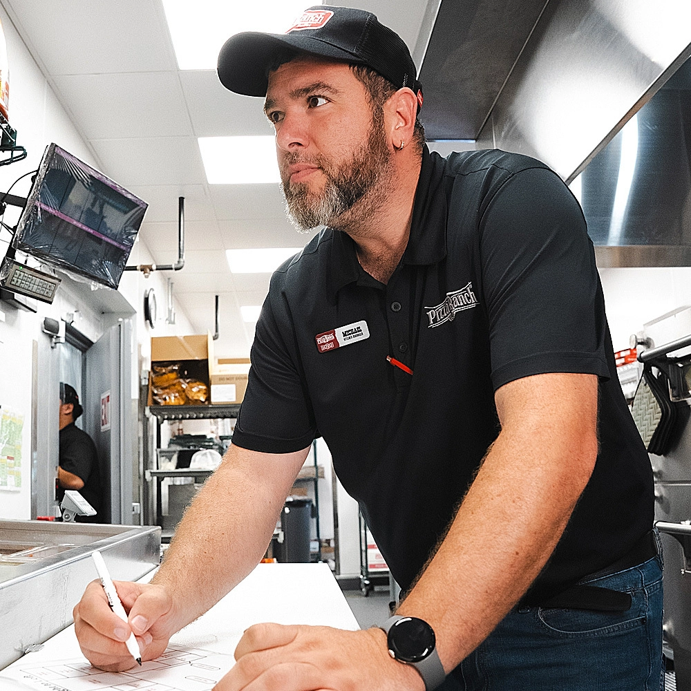 A man in a black uniform, wearing a black cap, writing on a paper at a fast-food restaurant, with equipment and supplies in the background.