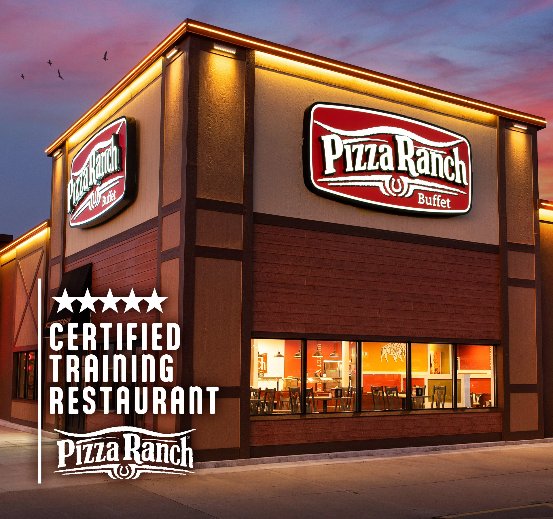 Two Pizza Ranch restaurant signs are mounted on a building with large windows showing the interior. The building is lit with warm exterior lighting during sunset.