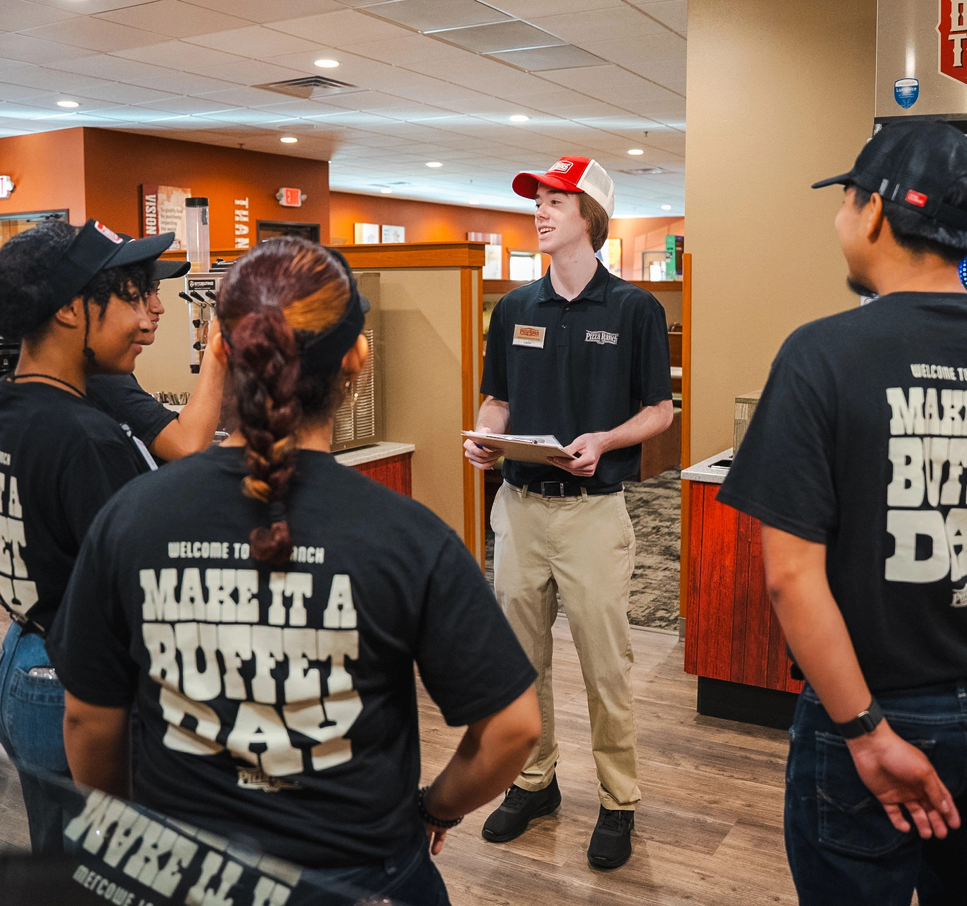 A group of employees gathered around a manager in a Pizza Ranch, wearing black shirts with "Make It a Buffet Day" printed on the back, listening to instructions.