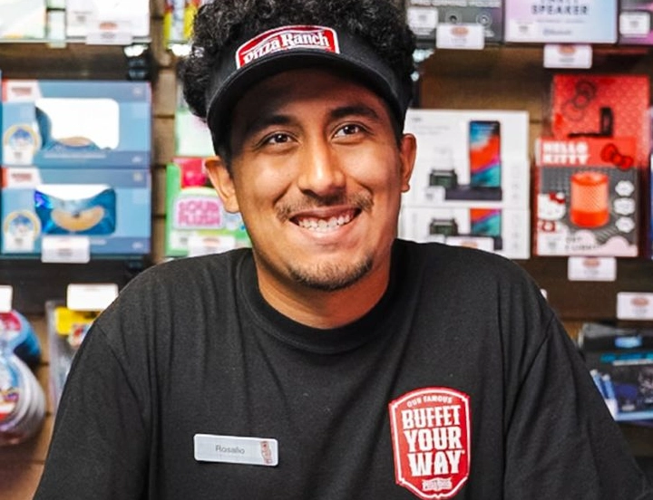 A smiling man wearing a black Pizza Ranch visor cap and a black T-shirt with a "Buffet Your Way" logo, sitting in front of shelves filled with various product boxes.