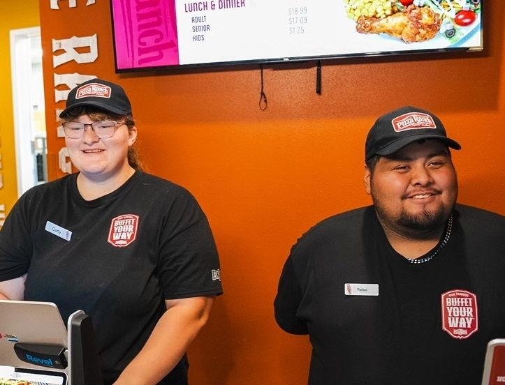 Two employees wearing black uniforms and caps with the Pizza Ranch logo stand behind the counter, smiling in front of an orange wall and digital menu screen.