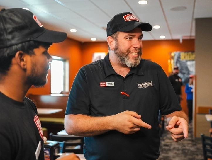 A man in a Pizza Ranch uniform, including a cap and polo shirt, smiling and gesturing with his hands while talking to a colleague in a restaurant setting.