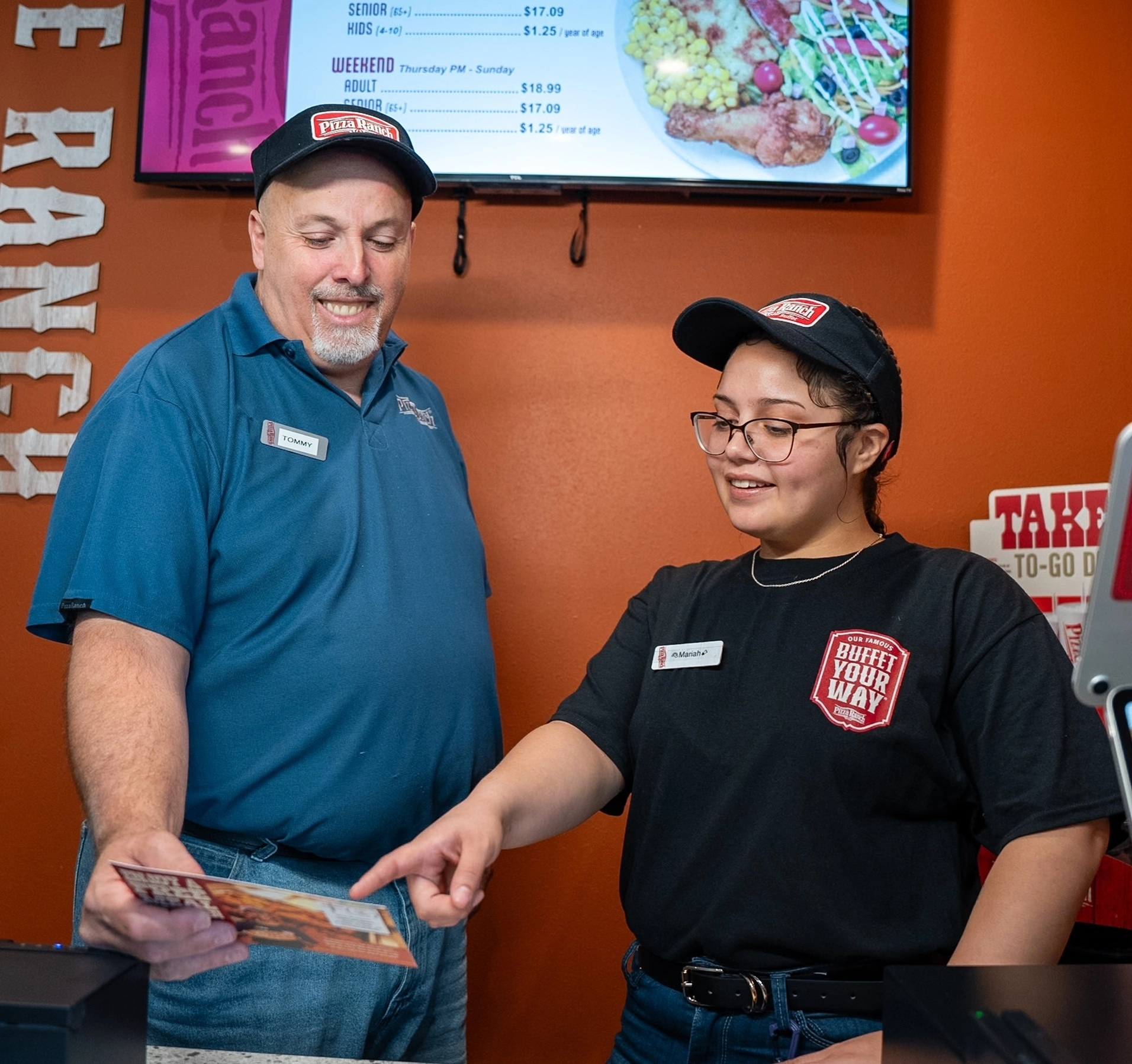 A man and a woman wearing Pizza Ranch uniforms and hats are smiling at each other while reviewing a menu or flyer inside a restaurant with an orange wall and a digital screen behind them.