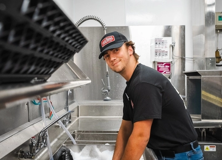 A young man in a Pizza Ranch uniform, wearing a cap and black shirt, washing dishes in a commercial kitchen with stainless steel sinks and equipment.