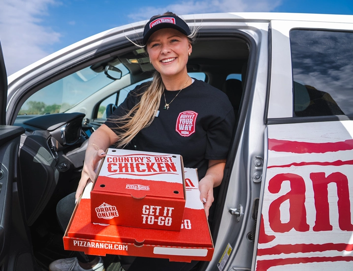 A smiling woman in a Pizza Ranch uniform and cap is sitting in a vehicle, holding a pizza box with a "Country's Best Chicken" label, ready for delivery.