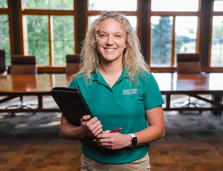A woman with curly blonde hair smiling and holding a clipboard and pen, wearing a teal Pizza Ranch uniform, standing in a conference room with large windows.