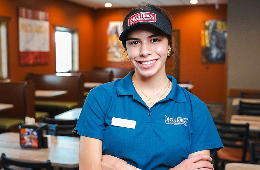 Smiling young woman in a blue Pizza Ranch uniform and black visor hat, standing with arms crossed inside a restaurant with orange walls.