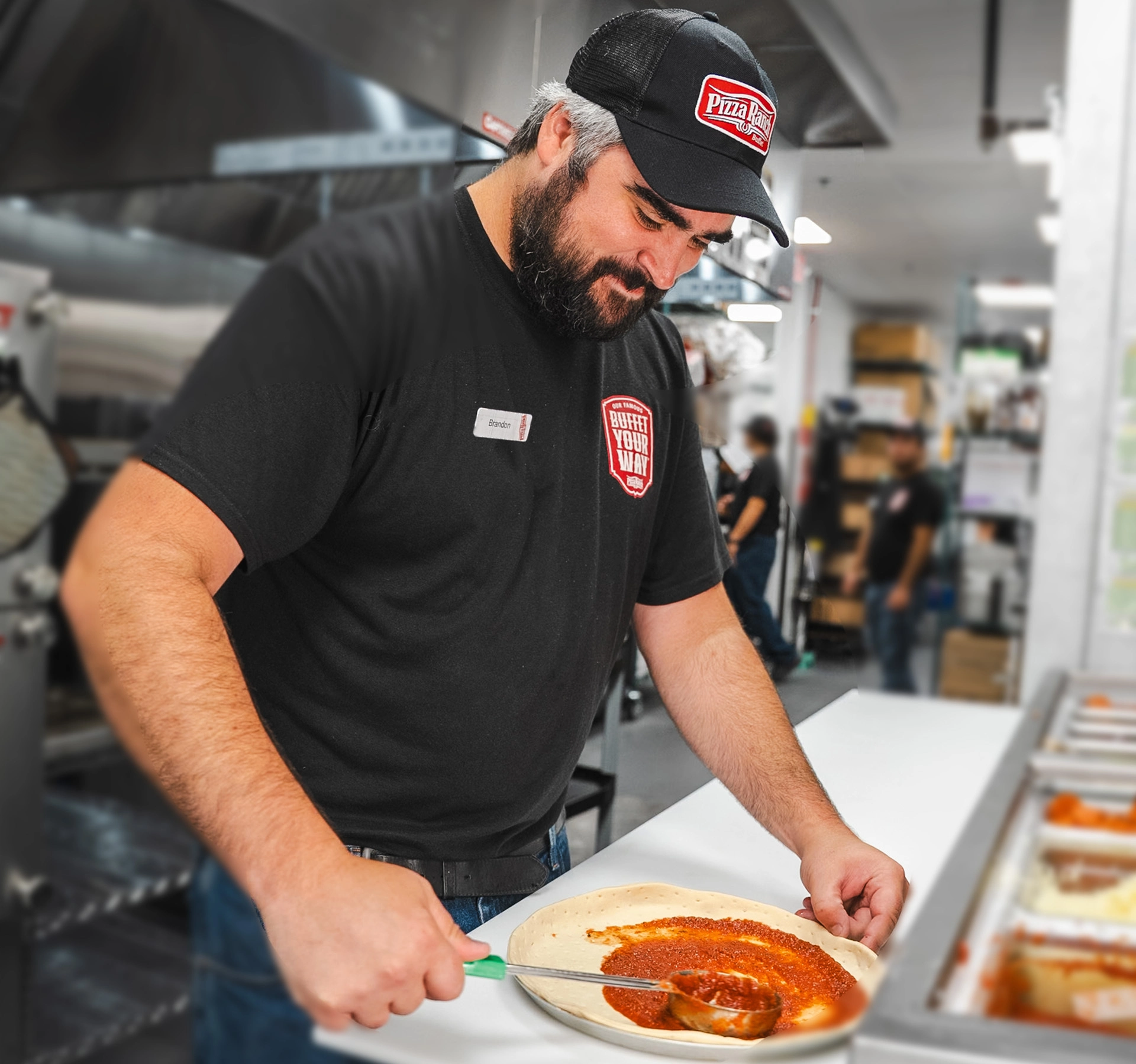 A man with a beard wearing a black cloth and cap with Pizza Ranch logo, spreading pizza sauce on dough at a restaurant kitchen, smiling while preparing pizza.