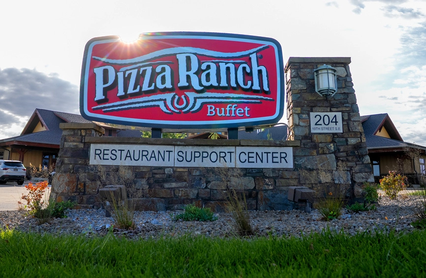 A large sign for Pizza Ranch Buffet mounted on a stone structure, with a smaller sign below indicating a restaurant support center; building and cars in background.