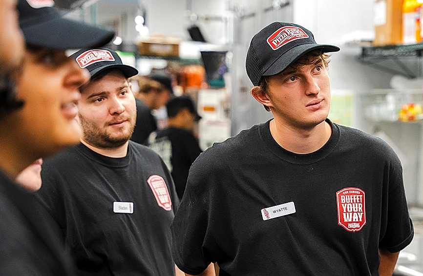 Three employees in black uniforms and caps with Pizza Ranch logos, standing in a kitchen area, appear to be listening attentively.