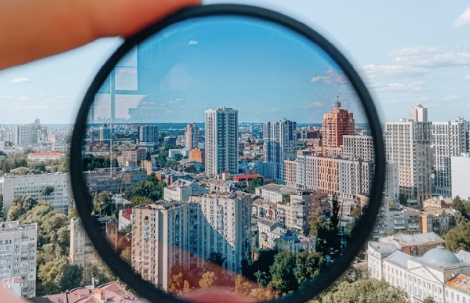 A hand holds a circular lens or filter in front of a cityscape, creating a fisheye view of tall buildings and trees under a mostly clear blue sky.